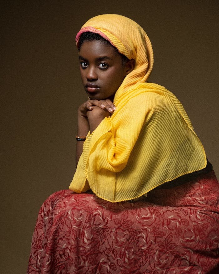 Portrait of a woman in a yellow hijab and red patterned dress looking thoughtfully.